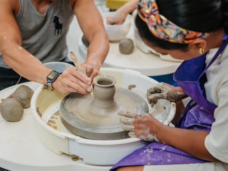 Two pairs of messy hands work on a pot together in a wheel throwing class.